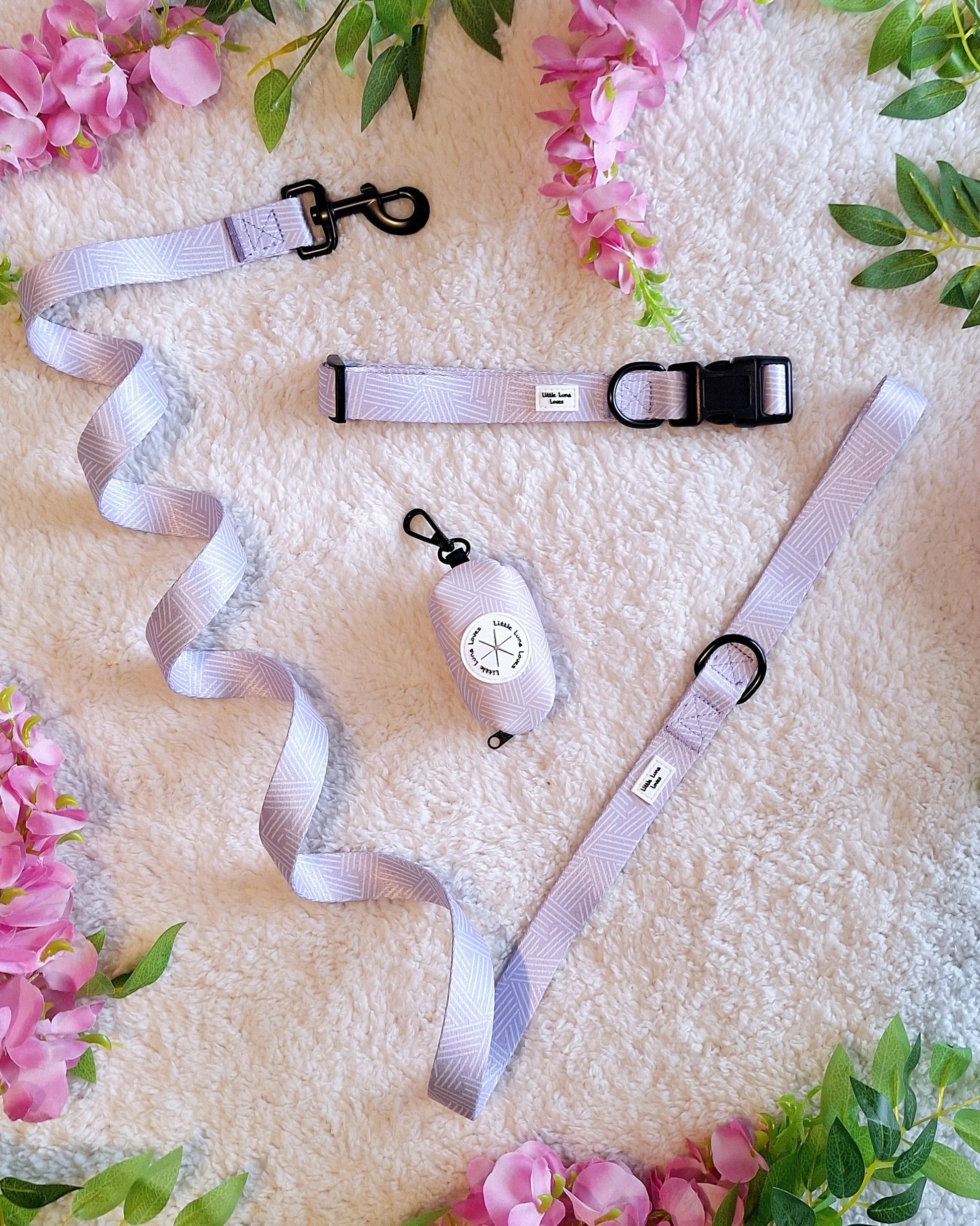 A dog lead and accessories displayed on a white surface surrounded by pink flowers, the dog lead and accessories are primarily gray and black.