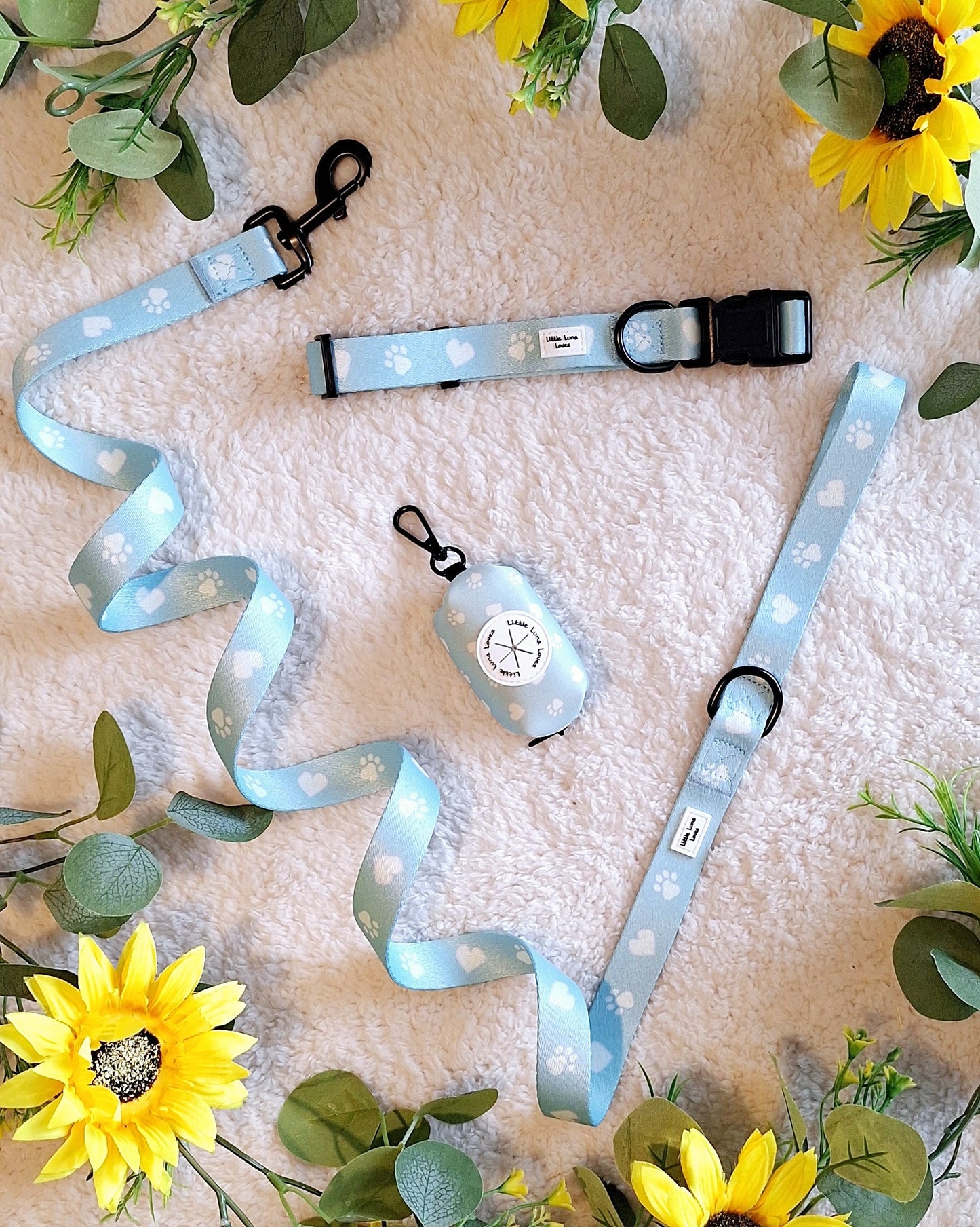 A dog lead with a light blue color and white paw print pattern, displayed on a white surface surrounded by yellow flowers and green leaves.