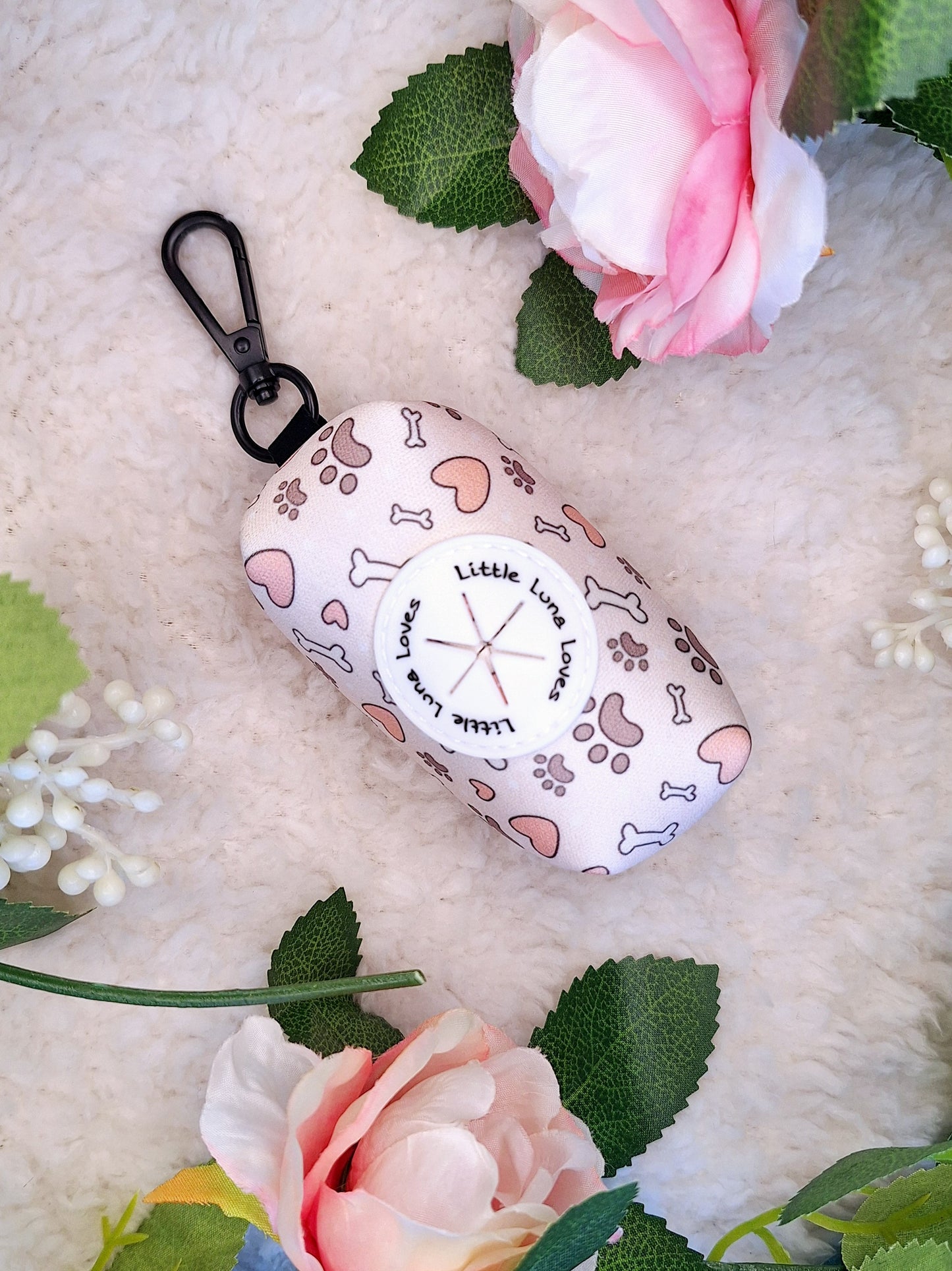 A neoprene fabric poop bag holder with a clip, featuring a white background with a pattern of bones and hearts, displayed on a faux fur surface with pink flowers around it.