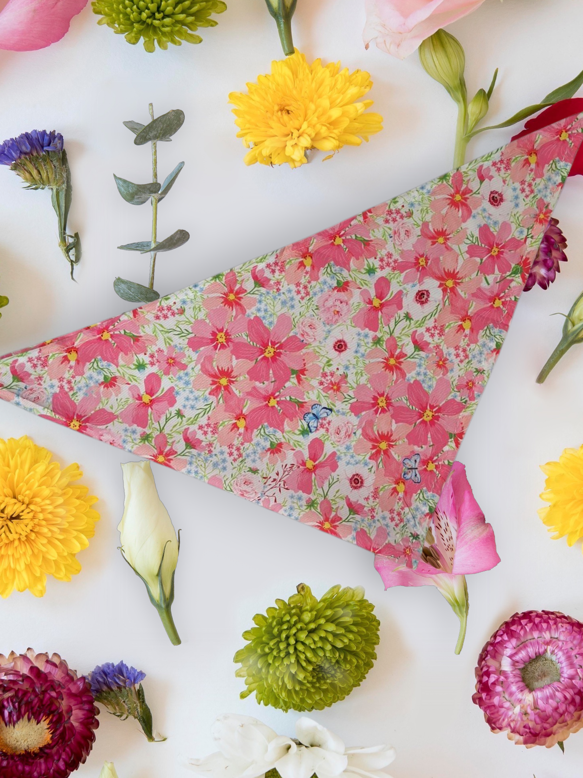 A floral patterned neck tie Dog bandana laid out flat with various colourful flowers arranged around it.