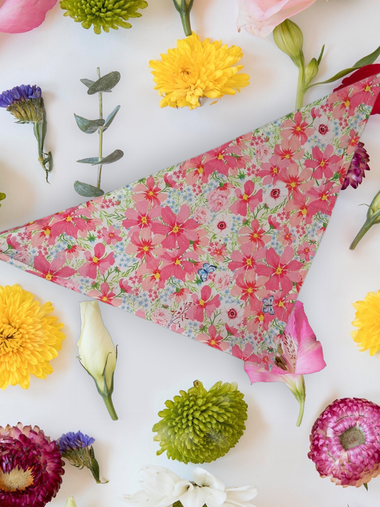 A floral patterned neck tie Dog bandana laid out flat with various colourful flowers arranged around it.