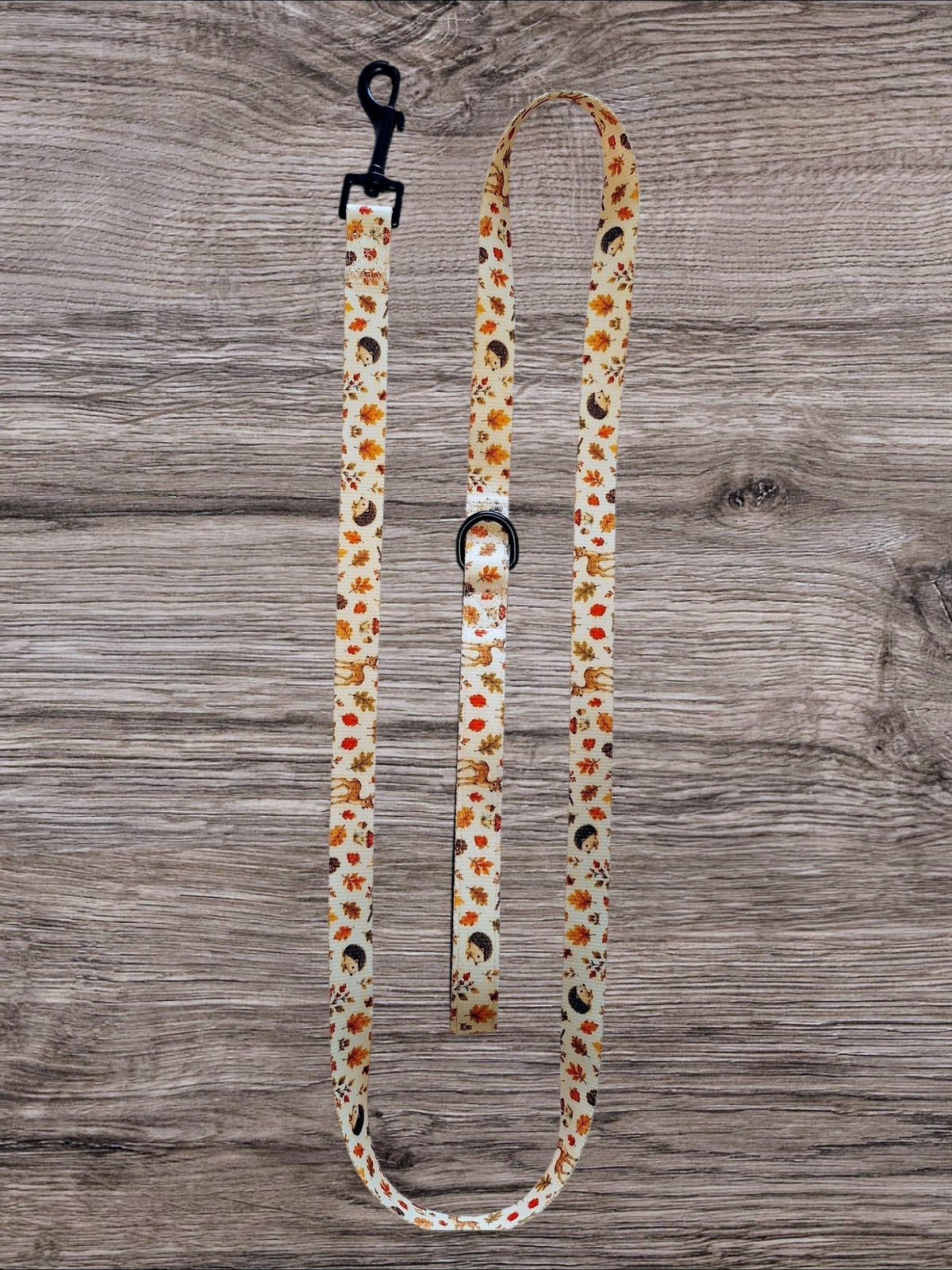 A fabric dog leash with a black metal clasp and matching buckle, featuring a cream background with a pattern of orange flowers, brown leaves, and black birds, laid flat on a gray wood plank surface.