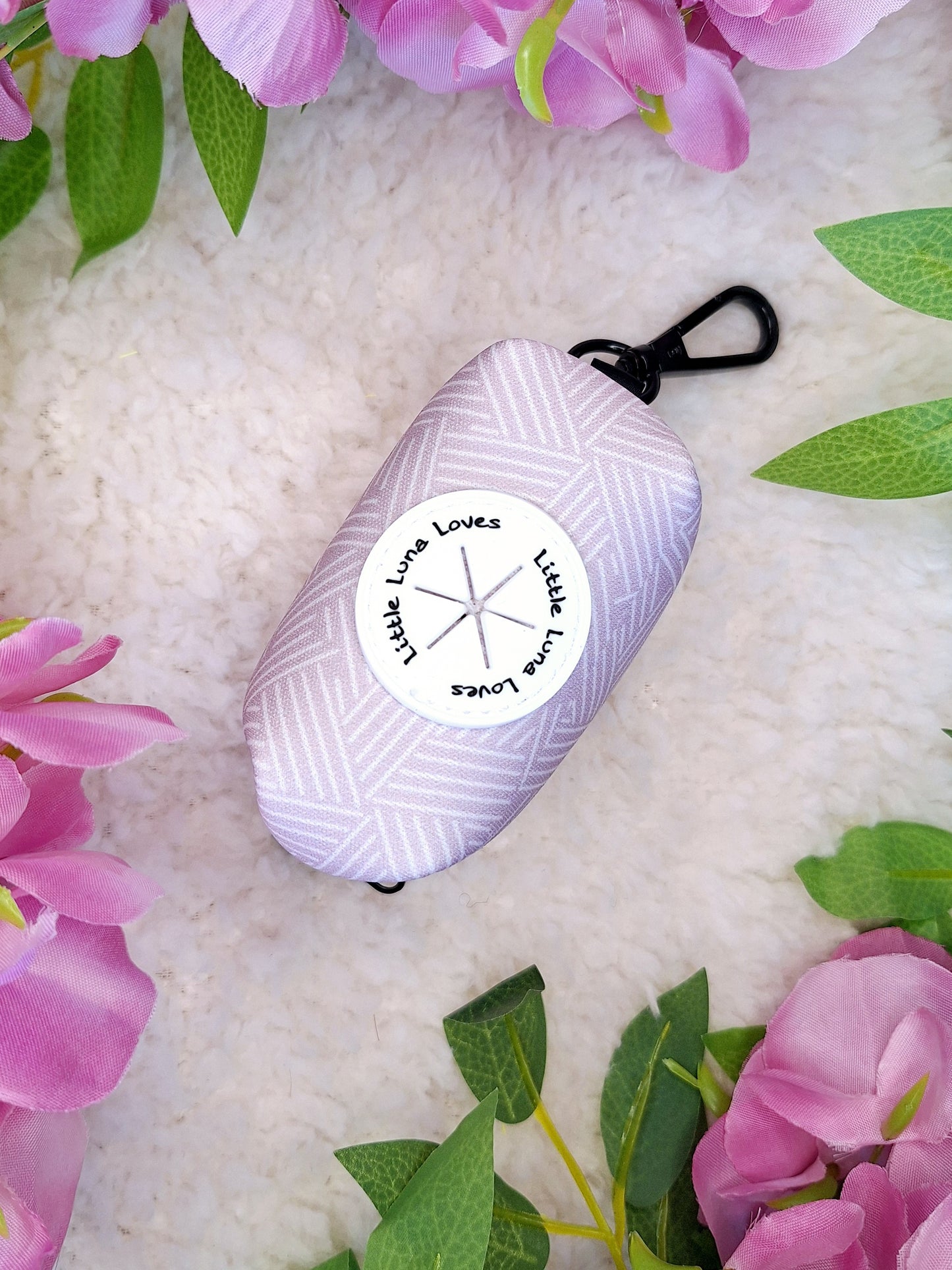 A stylish lavender and white striped pet waste bag holder with a clip and zip top, displayed on a fluffy surface surrounded by lavender-colored flowers.