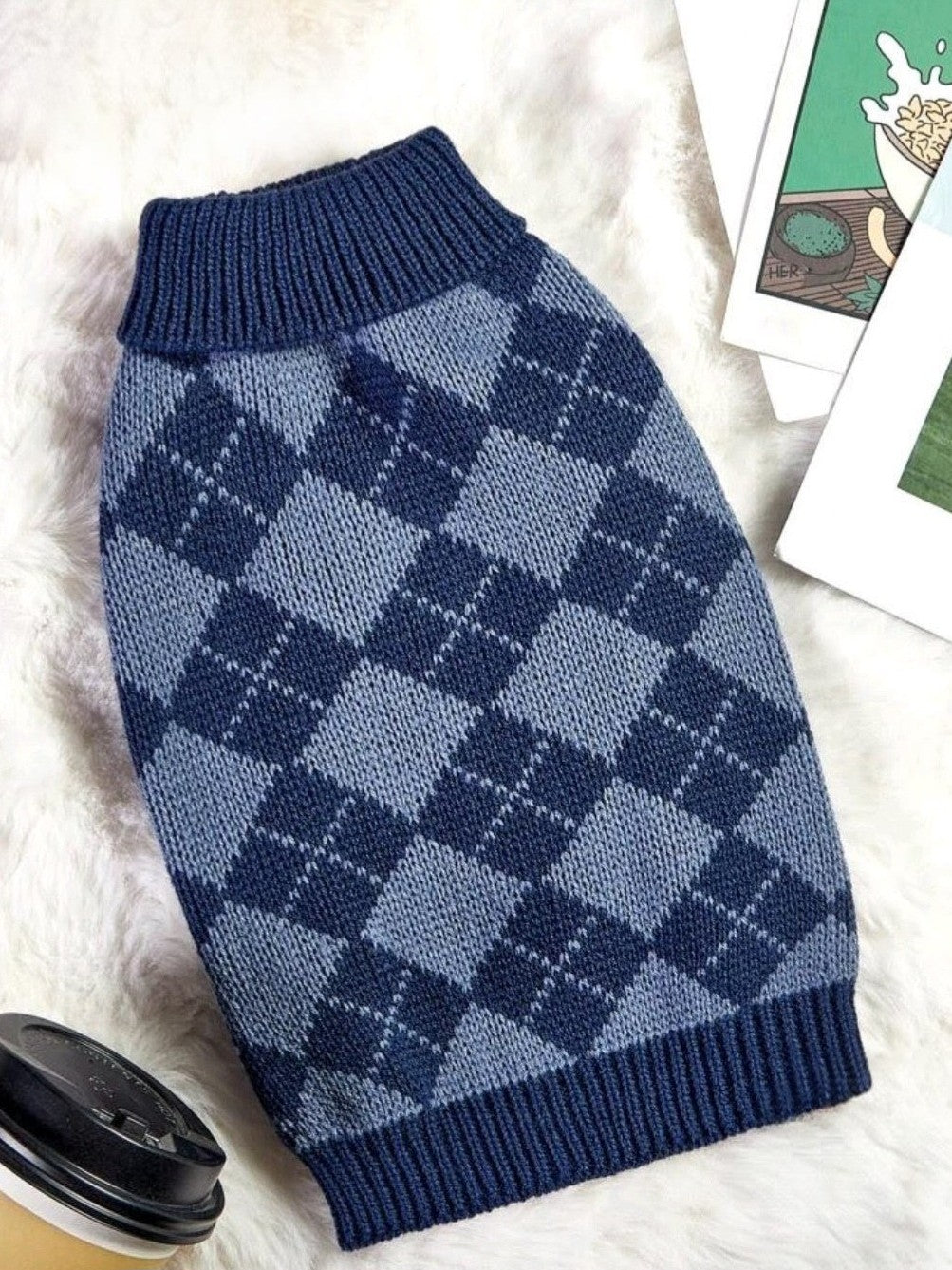 Blue checkered dog sweater on a white surface with a coffee cup and books in the background.