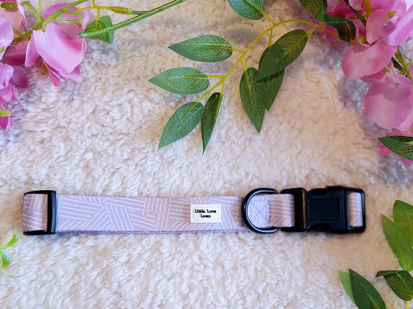 Adjustable dog collar in lavender and white colors with a quick release buckle and a D-ring, displayed on a fluffy surface with pink flowers around.