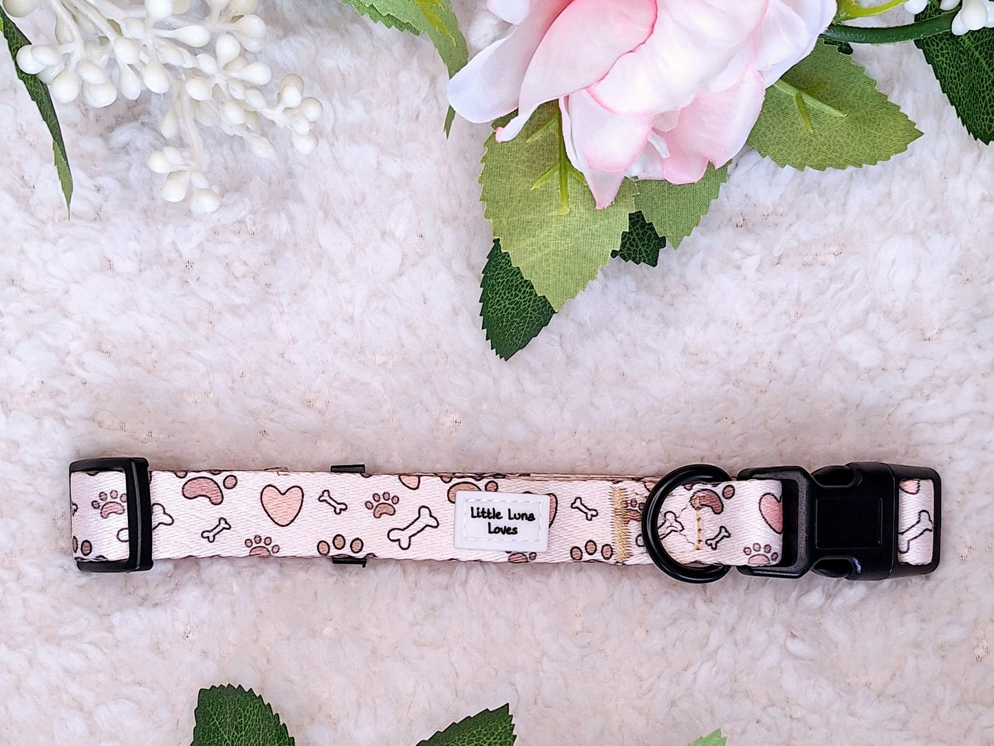A pink dog collar with white bones and hearts pattern, displayed on a white fluffy surface with a pink flower at the top left corner.