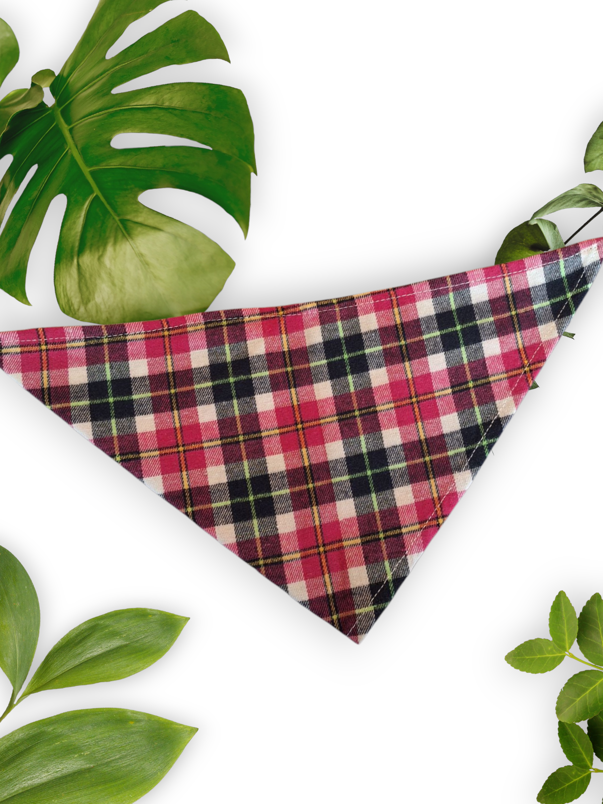 A pink plaid neck tie bandana for dogs placed on a white background surrounded by green leaves.