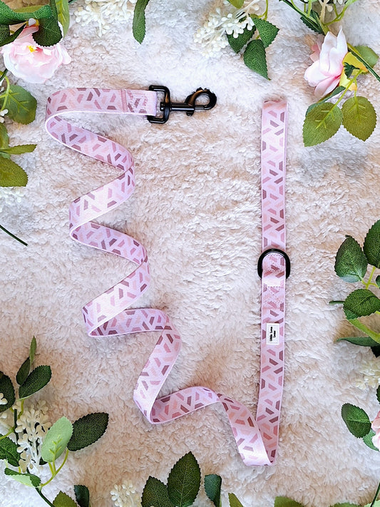A pink dog lead with a paw print pattern displayed on a white surface surrounded by green leaves and flowers.