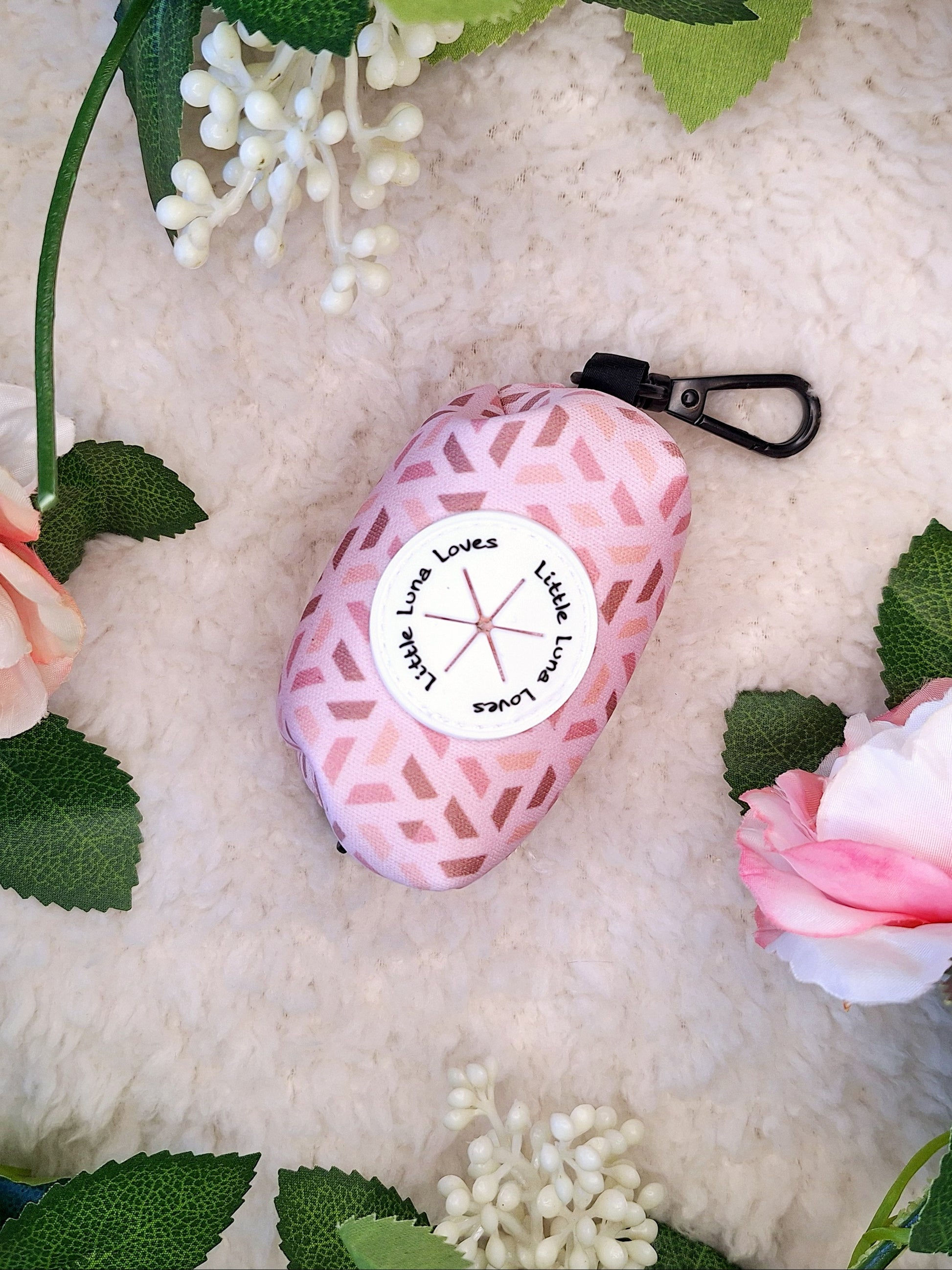 A pink neoprene poop bag holder with a clip, placed on a white surface surrounded by decorative flowers.