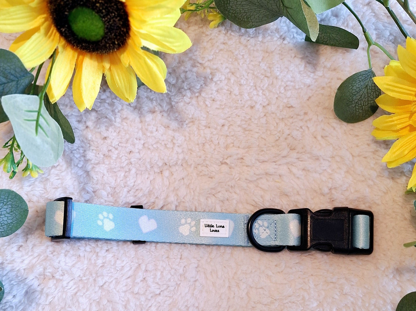 Adjustable blue dog collar with white paw prints and text that says 'Paws For Love', featuring a plastic buckle and D-ring, displayed on a fluffy background with sunflowers.