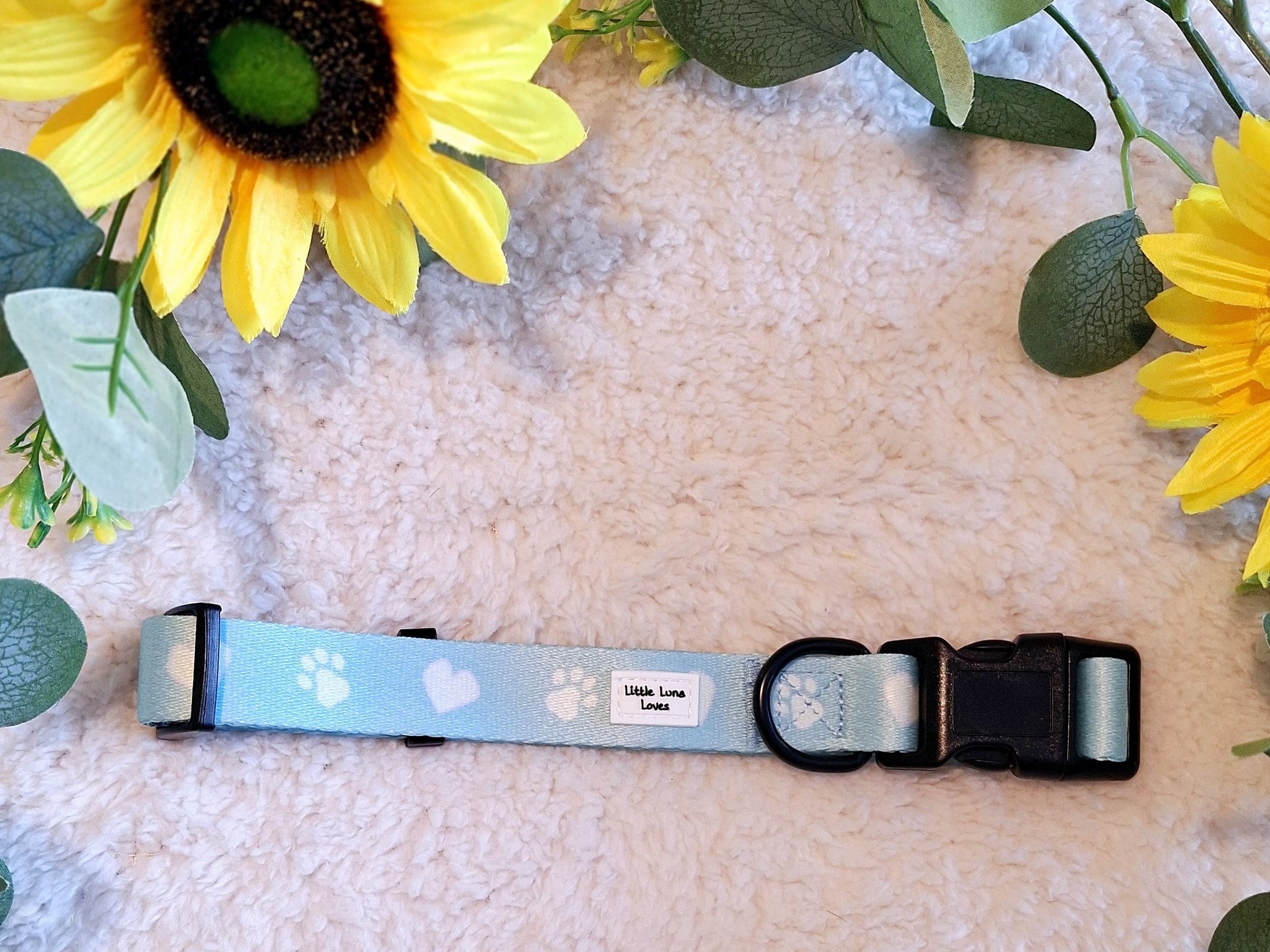 Adjustable blue dog collar with white paw prints and text that says 'Paws For Love', featuring a plastic buckle and D-ring, displayed on a fluffy background with sunflowers.