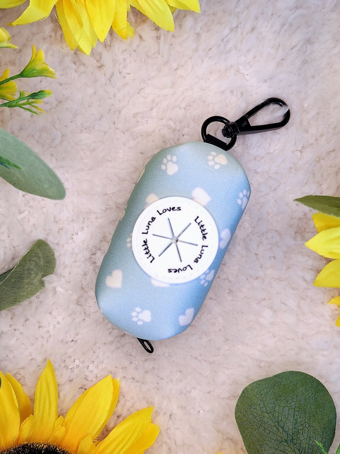 A blue neoprene poop bag holder with a clip, decorated with a white paw print and heart design, displayed on a white surface surrounded by sunflowers and leaves.