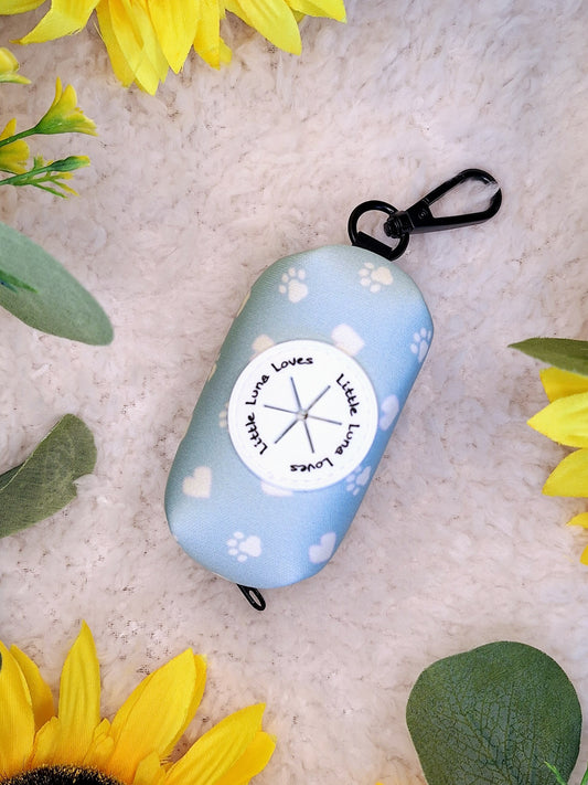 A blue neoprene poop bag holder with a clip, decorated with a white paw print and heart design, displayed on a white surface surrounded by sunflowers and leaves.