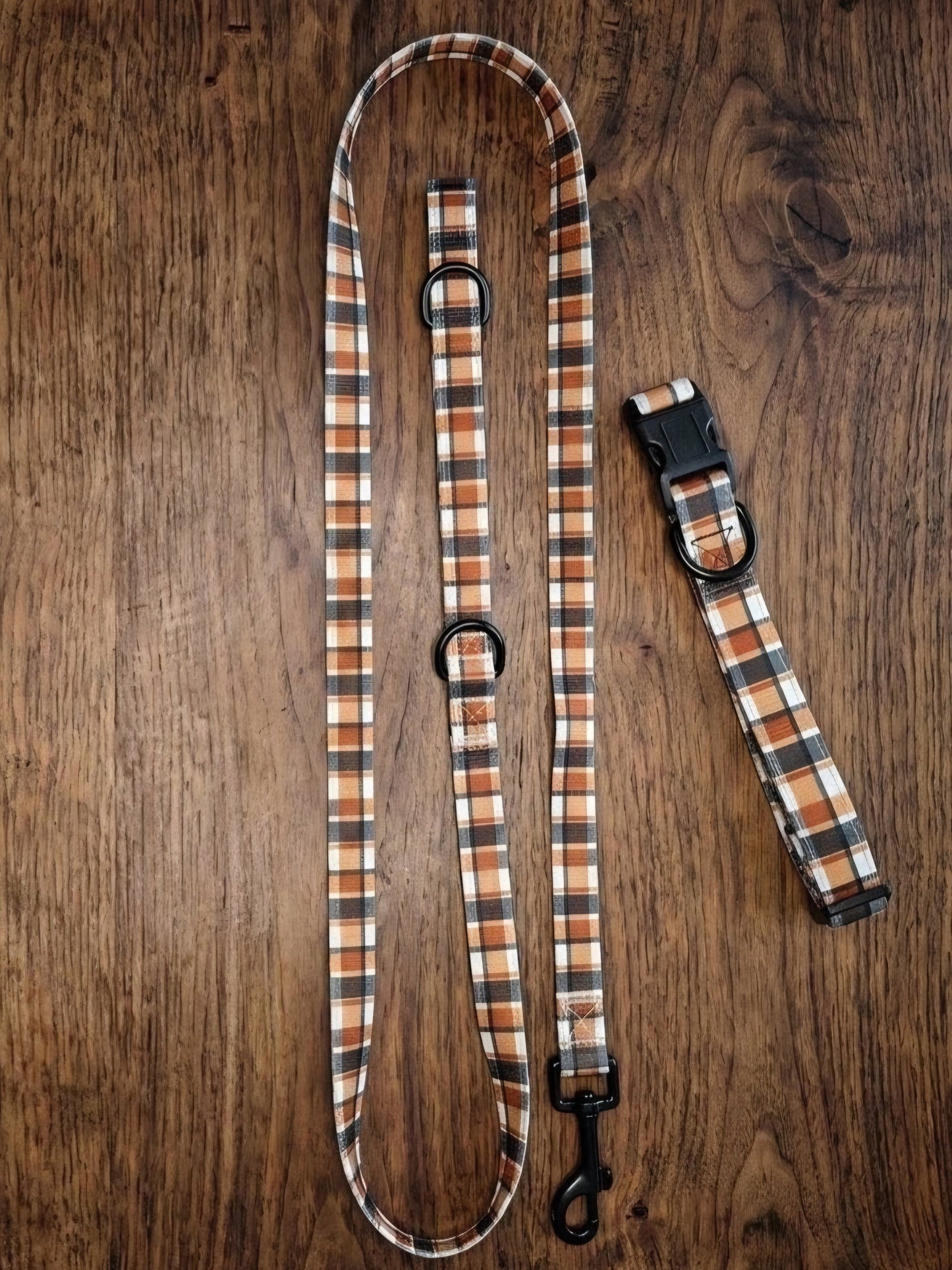 A dog leash and matching collar laid flat on a dark brown wood surface. The fabric has a plaid pattern in tan, brown, black, and white, with black metal hardware including rings, a buckle, and a snap hook.