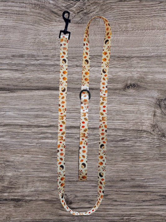 A fabric dog leash with a black metal clasp and matching buckle, featuring a cream background with a pattern of orange flowers, brown leaves, and black birds, laid flat on a gray wood plank surface.
