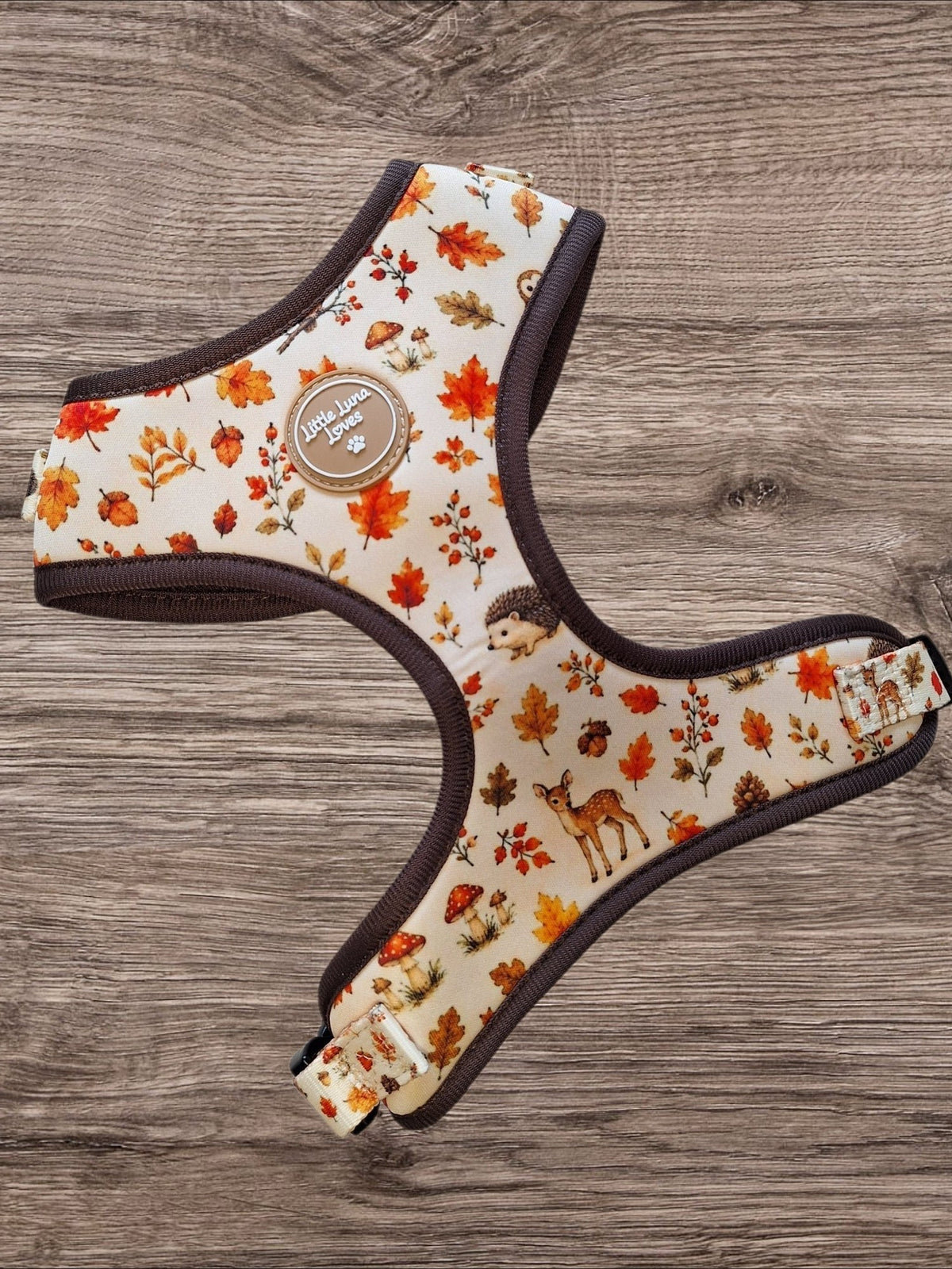 A dog harness with an autumn-themed print of orange and red leaves, pinecones, and deer on a cream background, featuring dark brown trim, laid flat on a gray-brown wooden surface.