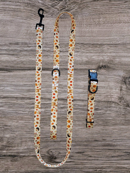 A dog leash and matching collar laid flat on a gray wood plank background. The fabric has a cream base with a repeating pattern of orange pumpkins, small red berries, black cat silhouettes, and white paw prints, with black plastic hardware.