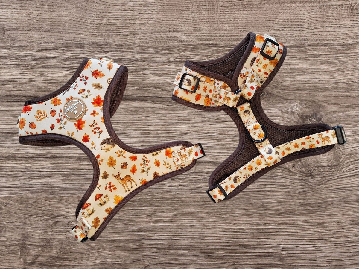 Two dog harnesses with a fall-themed print of leaves, acorns, and deer on a cream background, laid flat on a gray wood-textured surface.