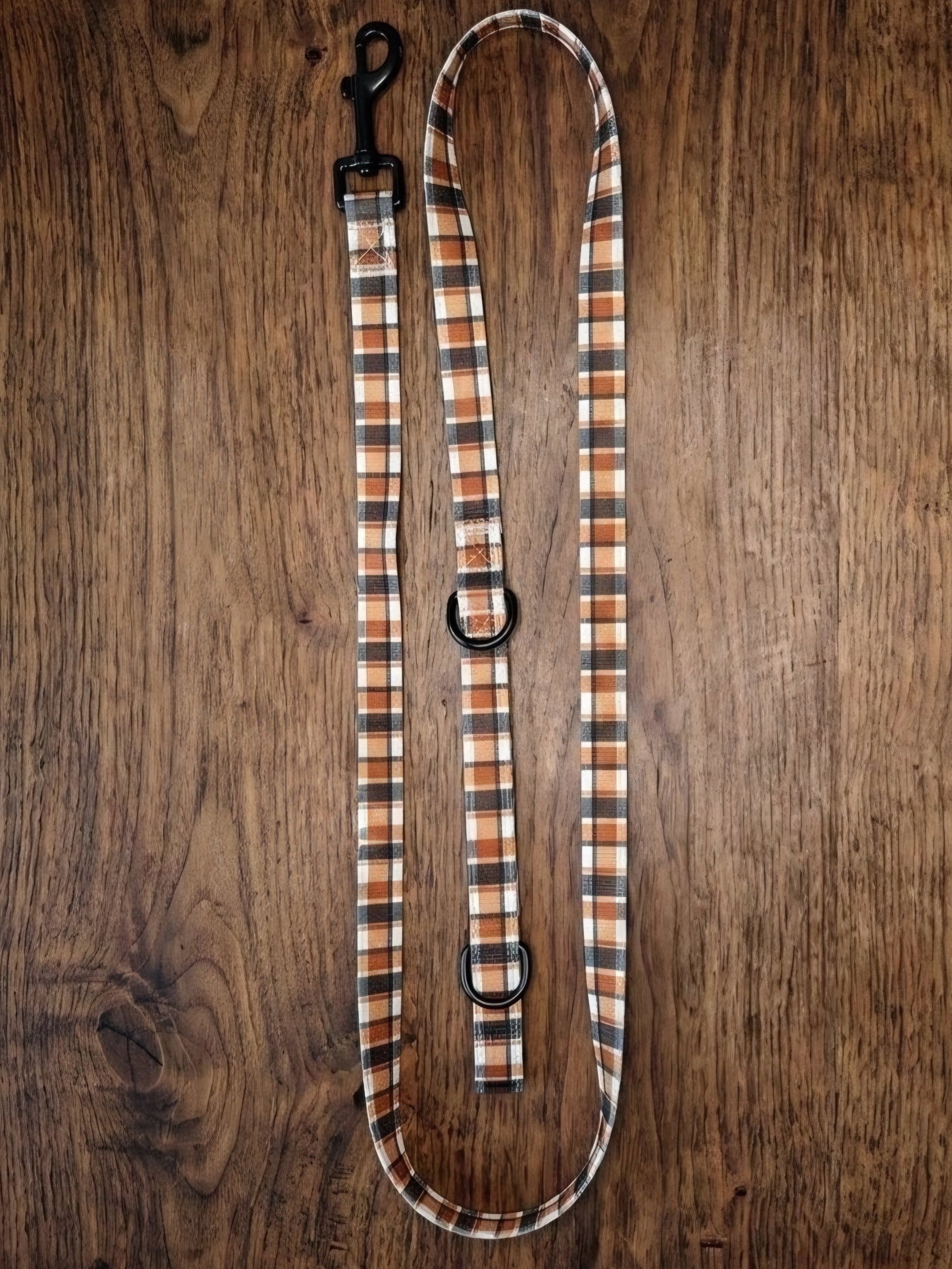 A plaid dog leash with a black metal clip and D-rings, laid flat on a dark brown wooden surface.