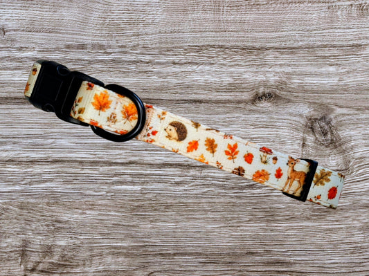 A fabric dog collar with a black plastic buckle and D-ring, featuring an autumn leaf and hedgehog pattern on a light cream background, laid flat on a gray wood-grain surface.
