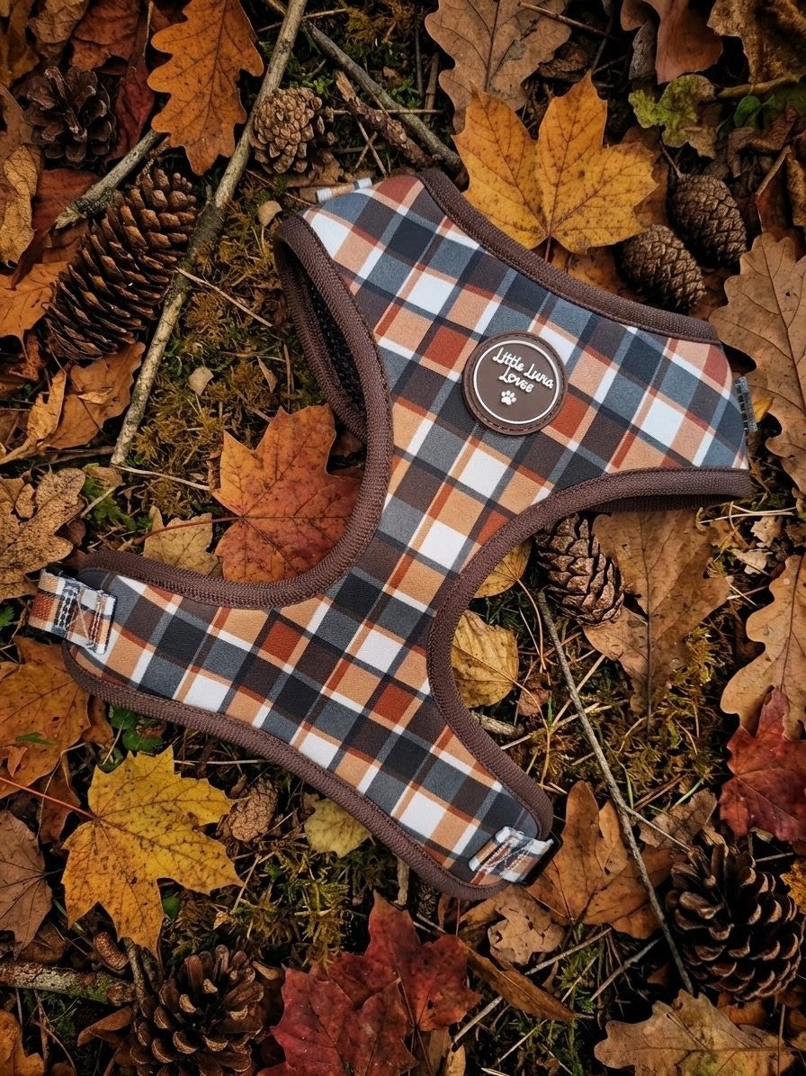 A plaid dog harness in brown, orange, black, and white, laid flat on a bed of autumn leaves and pine cones. The harness features a circular brown leather patch on the chest with the brand name 'Love Your Life'.