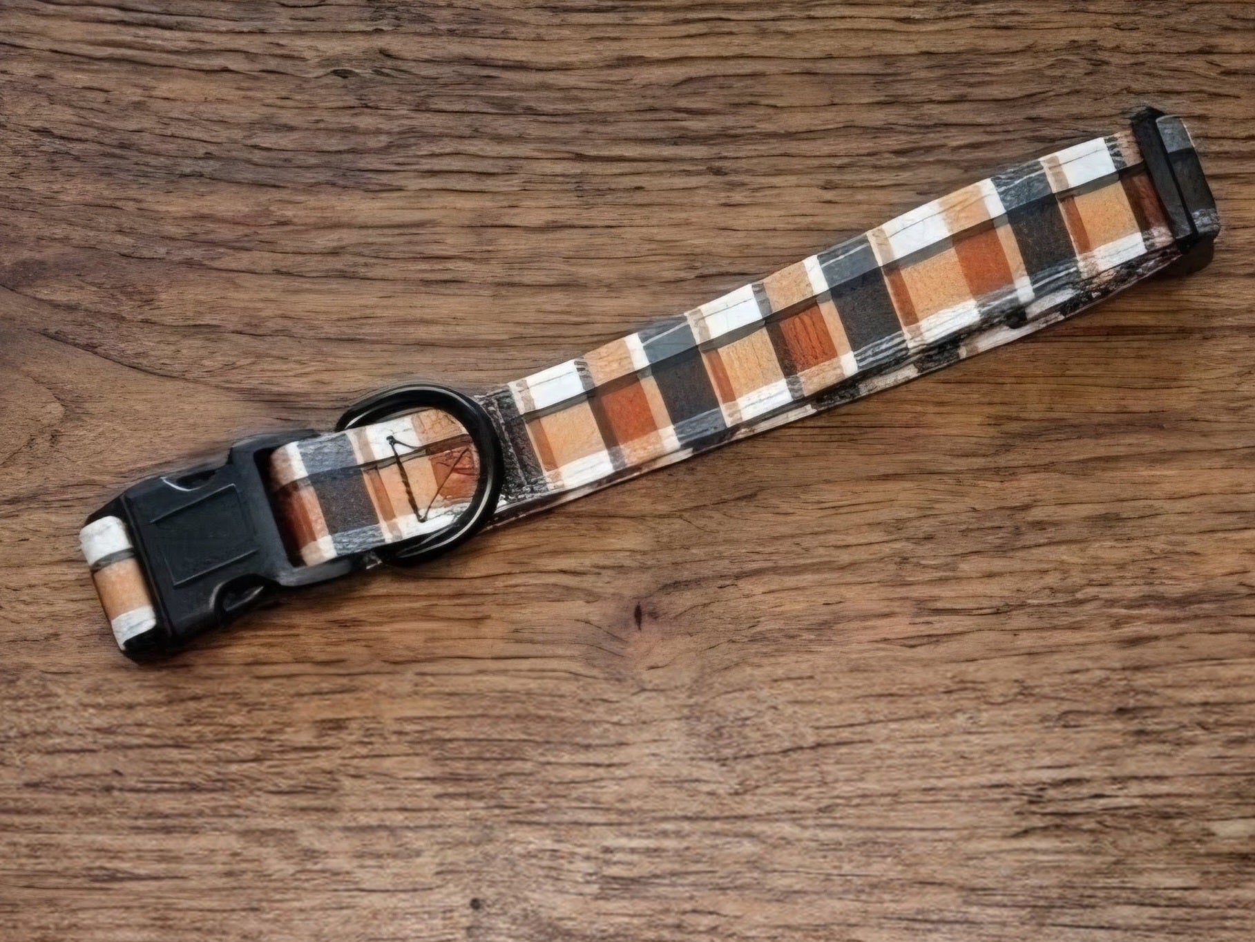 A plaid dog collar with black plastic hardware, featuring a pattern of brown, orange, black, and white stripes, displayed on a wooden surface.